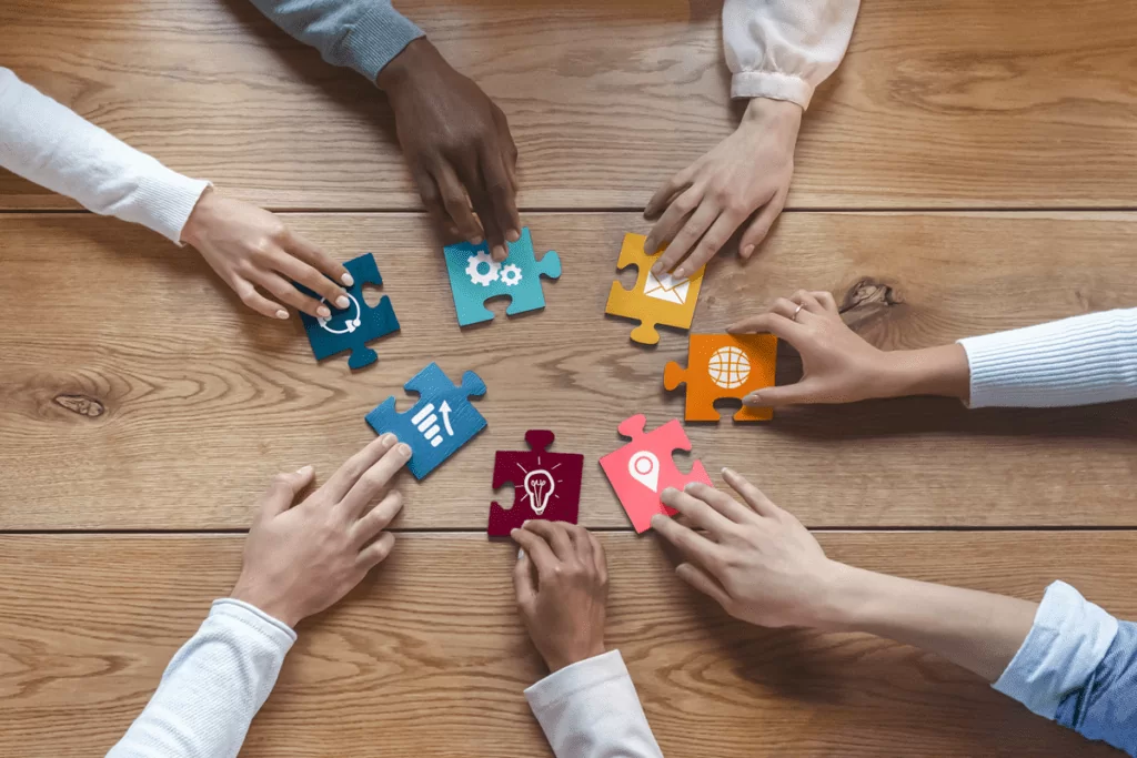Top view of diverse team members collaborating by connecting colorful puzzle pieces on a wooden table, symbolizing teamwork, collaboration, and problem-solving.