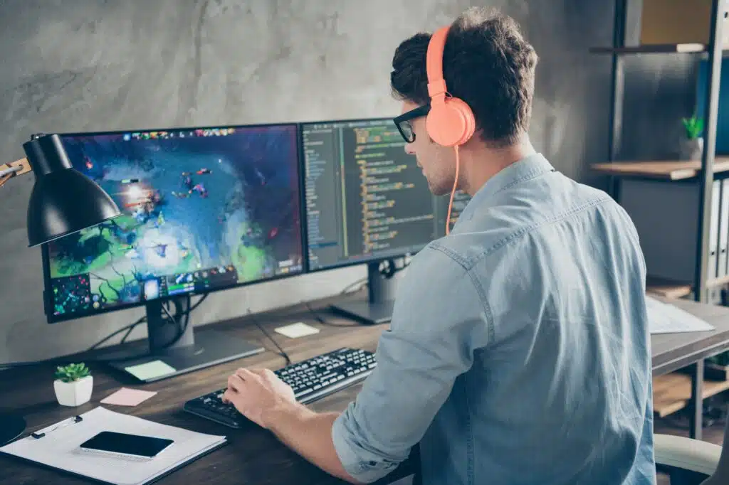 Game developer wearing headphones working at a dual-monitor setup, coding on one screen and testing a video game on the other